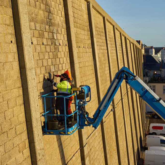 Two men on a blue boom arm make repairs to a tall cliff wall